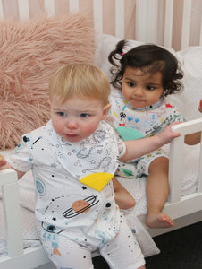 Babies sit in cot, wearing patterned baby clothes.