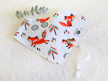 Load image into Gallery viewer, White bib with brilliantly bright orange foxes running and sitting, with tree-like pattern in black, orange and teal. White silky material coming out of bib to attach dummy/ comforter to. 