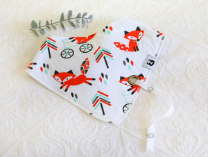 White bib with brilliantly bright orange foxes running and sitting, with tree-like pattern in black, orange and teal. White silky material coming out of bib to attach dummy/ comforter to. 