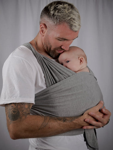 Image of a man holding a baby, with a charcoal grey baby carrier supporting the baby against the mans chest. 
